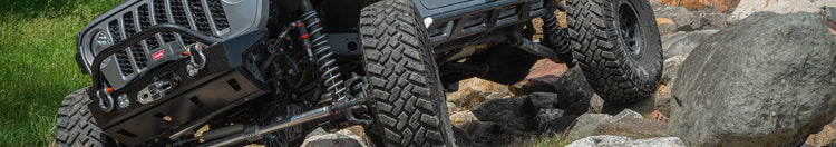 A dark gray custom off-road Jeep Wrangler is shown climbing over large rocks in an outdoor forest setting. The vehicle features custom graphics on the doors and hood, a front bumper guard, and large knobby tires.