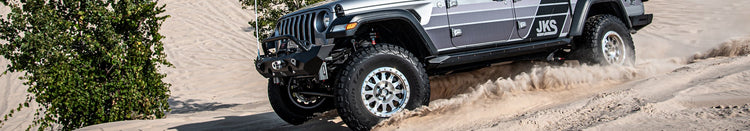 A silver Jeep Gladiator pickup truck with off-road modifications, including a front bumper mount and a rear winch, driving up a sandy dune in a beach environment.